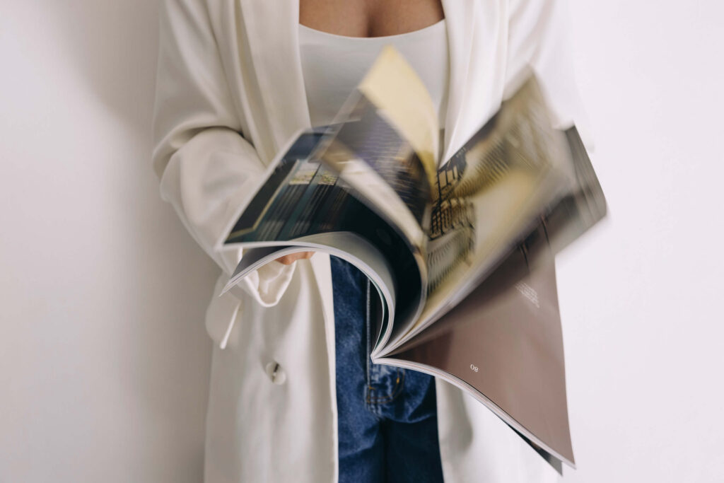 A woman in a white blazer and blue jeans flips through a glossy magazine. The motion blur suggests dynamic browsing, evoking interest and curiosity.