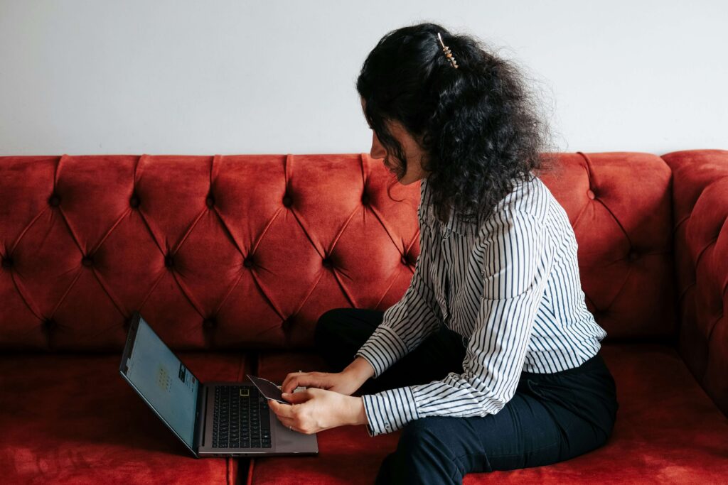 Woman sitting on a red sofa using a laptop while holding a credit card.